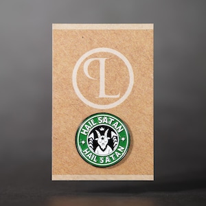 May include: A circular pin badge featuring a goat head design within a green and white border, with the words "HAIL SATAN". The badge is on a brown card with a white "L" and circle design.