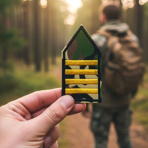 May include: A military rank insignia patch held in front of a person in a forest. The patch has a camouflage background with four yellow stripes. The person in the background is wearing camouflage clothing and a backpack.