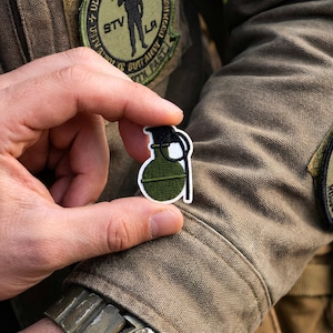 May include: A small, embroidered patch depicting a green hand grenade with a black top and white detailing. The patch is held between fingers against a tan military jacket, highlighting the patch's design and detail.
