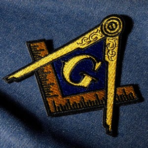 May include: Embroidered patch featuring a Masonic square and compass design in gold and blue. The patch includes a gold letter "G" in the center, set against a blue background, and is set on a blue denim fabric.