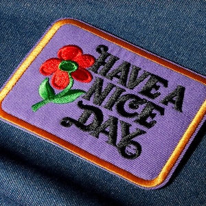 Peut inclure: Un écusson rectangulaire violet brodé avec une bordure dorée et les mots "HAVE A NICE DAY" en noir. Une fleur rouge avec des feuilles vertes est sur le côté gauche. L'écusson est sur un fond en denim bleu.