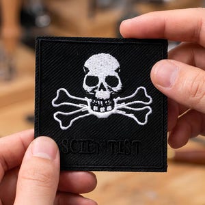 May include: A black square patch featuring a white skull and crossbones emblem. The word "SCIENTIST" is embroidered in white beneath the skull. The patch is held by two hands.