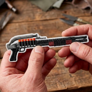 May include: An embroidered patch depicting a shotgun in black and white, with red and orange bullet details. The patch is held in a person's hands. The background is a wooden surface.