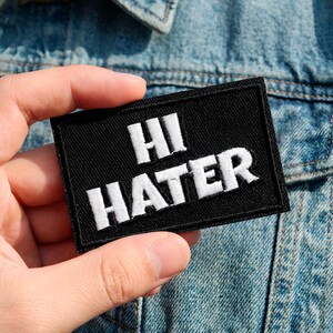 May include: A black rectangular patch with the words "HI HATER" embroidered in white. The patch is held in front of a denim jacket.