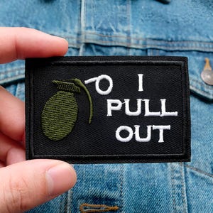 Könnte beinhalten: Schwarzer rechteckiger Aufnäher mit weißem Text "I PULL OUT". Der Aufnäher zeigt ein gesticktes grünes Granaten-Design. Der Aufnäher wird vor einer Jeansjacke gehalten.