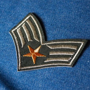 May include: Embroidered patch featuring a star and stripes design in shades of green, tan, and orange. The patch is set against a blue denim background. The patch has a military or vintage aesthetic.