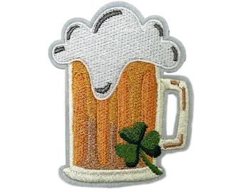 以下が含まれることがあります： 泡立った泡と緑のクローバーが付いたビールジョッキの刺繍パッチ。ジョッキは琥珀色のビールで満たされ、白いハンドルが付いています。パッチは灰色の縁取りで、衣類やアクセサリーに適しています。