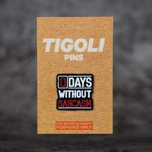 Op de afbeelding: Een rechthoekige pin met de tekst "0 DAYS WITHOUT SARCASM" in wit en rood op een zwarte achtergrond. De pin zit op een bruine kaart met de woorden "TIGOLI" en "PINS" in het wit gedrukt. De onderkant van de kaart luidt "FOR ENTERTAINMENT PURPOSES ONLY."