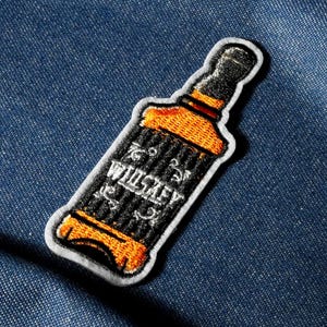 May include: Embroidered patch in the shape of a whiskey bottle. The patch features a black and orange design with the word "Whiskey" in white lettering. The patch has a white border and is set against a blue denim background.