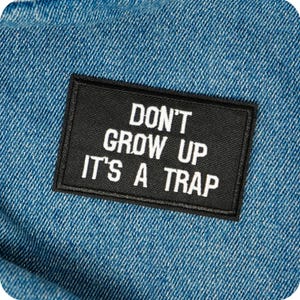 May include: A black rectangular patch with white text reading "DON'T GROW UP IT'S A TRAP" is sewn onto blue denim. The patch has a black border and the denim has a textured appearance.
