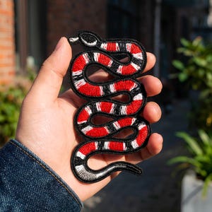 Può includere: Toppa ricamata a forma di serpente, nera, rossa e bianca. Il design del serpente è dettagliato con un contorno nero, strisce rosse e bianche e una forma curva. La toppa è tenuta in mano.