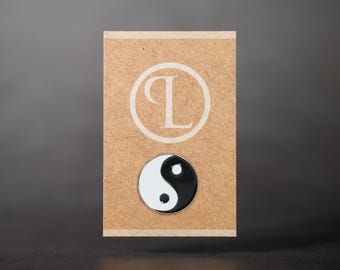 May include: A round yin and yang pin with a black and white design, set against a tan card. The card features a white circular logo with the letters "PL". The pin is a symbol of balance.