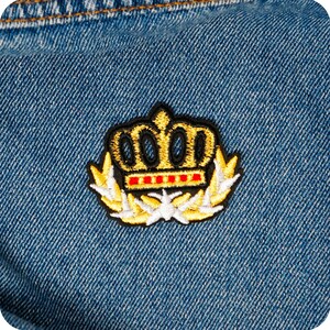 May include: Embroidered patch with a gold crown design, black outline, red details, and laurel wreath, sewn onto blue denim. The patch presents a classic, regal emblem, suitable for clothing or accessories.