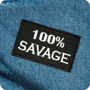 Könnte beinhalten: Nahaufnahme eines blauen Jeansstoffs mit einem schwarzen rechteckigen Aufnäher. Der Aufnäher zeigt den weißen Text "100% SAVAGE". Der Jeansstoff hat eine klassische blaue Farbe und eine strukturierte Optik.