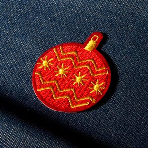May include: A red embroidered Christmas bauble patch with gold zigzag and starburst designs. The patch has a gold loop at the top, set against a blue denim background. Ideal for festive crafts and adding a touch of holiday cheer.
