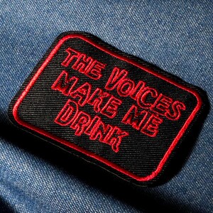 Op de afbeelding: Een zwarte patch met rode geborduurde tekst die luidt "THE VOICES MAKE ME DRINK". De patch heeft een rode rand en is geplaatst tegen een blauwe denim achtergrond. De patch is rechthoekig met afgeronde hoeken.