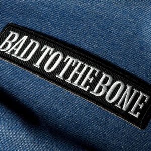 Könnte beinhalten: Ein schwarzer rechteckiger Aufnäher mit dem gestickten Text "BAD TO THE BONE" in Weiß. Der Aufnäher befindet sich auf einem blauen Jeansstoffhintergrund.