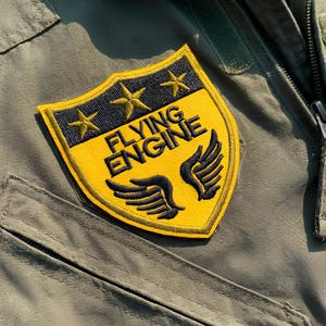 Può includere: Toppa ricamata gialla e nera con le parole "FLYING ENGINE" e due ali nere. La toppa presenta anche tre stelle dorate ed è cucita su un capo verde con cerniera e tasca.