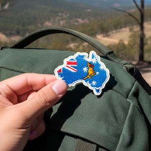 Könnte beinhalten: Ein blau-weißer, gestickter Aufnäher in Form von Australien, mit der australischen Flagge und einem Känguru. Der Aufnäher wird vor einem grünen Rucksack gehalten, mit einem verschwommenen Hintergrund im Freien.