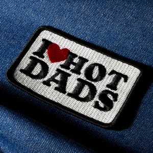 Puede incluir: Parche bordado con el texto "I ❤️ HOT DADS" en letras negras, un corazón rojo y un fondo blanco. El parche tiene un borde negro y está sobre un fondo de mezclilla azul.