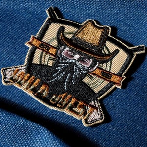 Könnte beinhalten: Gestickter Aufnäher mit einem Cowboy mit langem Bart und Hut. Der Aufnäher hat einen beigefarbenen Hintergrund mit einem schwarzen und braunen Cowboy-Design. Der Text "ESTD 1983" befindet sich auf einem Banner.