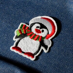 Op de afbeelding: Geborduurde pinguïn patch met een witte body, zwarte vleugels en een rood-groene sjaal. De pinguïn draagt een rood-witte hoed en heeft rode voeten. De patch is ontworpen om op stof te naaien of te strijken.
