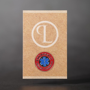 Può includere: Spilla rotonda con le parole "EMERGENCY MEDICAL TECHNICIAN" in lettere rosse. Il centro presenta un simbolo blu della stella della vita. La spilla è su uno sfondo beige con un logo bianco "P L".