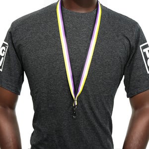 May include: A dark gray t-shirt with the letters "T" and "G" in white boxes on the sleeves. A non-binary pride flag ribbon is around the neck, with purple, white, and yellow stripes. A black clasp is at the end of the ribbon.