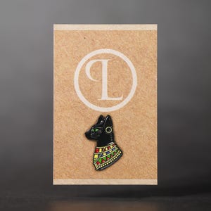 May include: A black cat pin with green eyes and a colourful collar, set against a tan card with a white circular logo. The pin is a stylized representation of an Egyptian cat deity.