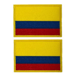 Puede incluir: Dos parches rectangulares con la bandera de Colombia. Cada parche tiene una sección superior amarilla, una sección media azul y una sección inferior roja. El diseño de la bandera está bordeado por un ribete dorado.