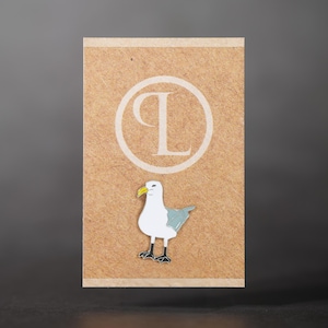 May include: A rectangular card with a brown background and a white circle with the letter "L" inside. A cartoon seagull with white, gray, yellow, and black details is at the bottom. The card is on a dark gray background.