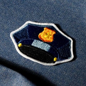 Puede incluir: Parche bordado de un gorro de policía. El parche azul marino tiene un borde blanco, un emblema dorado y una visera negra. El gorro tiene dos pequeños detalles amarillos. El parche está sobre un fondo de mezclilla azul.