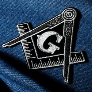 May include: Black and white embroidered patch featuring a Masonic square and compass design with the letter "G" in the center. The patch is on a blue denim background. The square and compass are outlined in white.