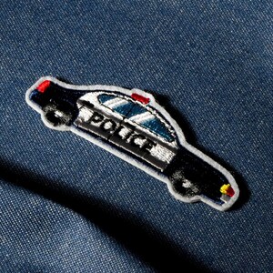 May include: Embroidered police car patch. The black car has white, red, and yellow details. The word "POLICE" is embroidered on the side. The patch is set against a blue denim fabric background.