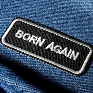 May include: A black rectangular patch with the words "BORN AGAIN" embroidered in white. The patch has a white border and is set against a blue denim background. It's likely an iron-on or sew-on accessory for clothing or bags.