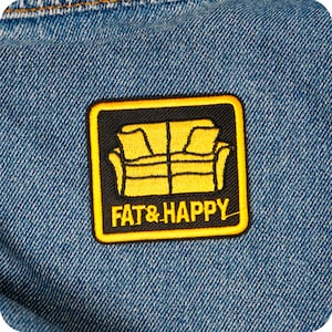 Può includere: Una toppa quadrata con bordo nero e design di divano giallo. La toppa ha le parole "FAT & HAPPY" in giallo. La toppa è cucita su un tessuto denim blu.