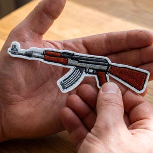 Könnte beinhalten: Gestickter Aufnäher in Form eines AK-47 Sturmgewehrs. Der Aufnäher hat einen grauen Lauf und Empfänger, einen rotbraunen Schaft und Handschutz sowie ein schwarzes Magazin. Der Aufnäher ist weiß umrandet.