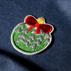 May include: Embroidered Christmas ornament patch with a green base, gray accents, and a red bow. The patch has a white border and a gold loop at the top. The patch is on a blue denim background.