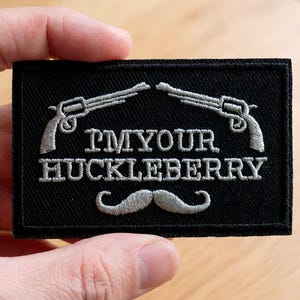 Peut inclure: Écusson rectangulaire noir avec des revolvers argentés brodés, une moustache et le texte "I'M YOUR HUCKLEBERRY". L'écusson est tenu dans la main d'une personne.
