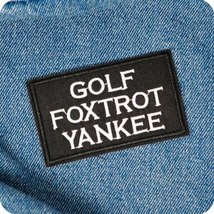 May include: A black rectangular patch with the words "GOLF FOXTROT YANKEE" in white lettering is sewn onto a blue denim fabric. The patch is centered and the text is in a bold, sans-serif font. The denim has a diagonal weave.