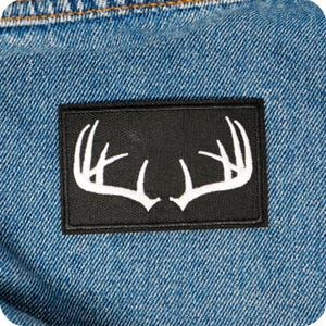 Puede incluir: Un parche rectangular negro con un diseño de cuernos blancos está cosido sobre una tela vaquera azul. El parche presenta un contorno detallado de cuernos de ciervo, creando un contraste visual con el fondo vaquero. La mezclilla tiene una apariencia clásica y texturizada.