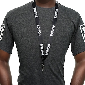 May include: A charcoal grey t-shirt with white text and a black lanyard. The lanyard is printed with the word "POLICE" in white. The sleeves of the shirt have white text and graphics. The shirt is made of a soft material.