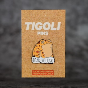 May include: A novelty pin featuring a cartoon toad with the text "TOAD YOU SO" on a white banner. The pin is on a brown card with the text "TIGOLI PINS" and "FOR ENTERTAINMENT PURPOSES ONLY". The toad is orange with brown spots.