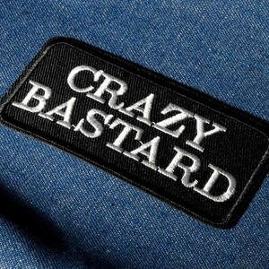 May include: A black rectangular patch with white embroidered text that reads "CRAZY BASTARD". The patch is set against a blue denim background. The text is in a bold, sans-serif font.