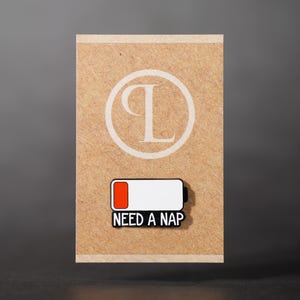 Può includere: Spilla rettangolare con un'icona della batteria quasi scarica, con la scritta nera "NEED A NAP". La spilla è su una carta marrone con un logo "P" bianco.