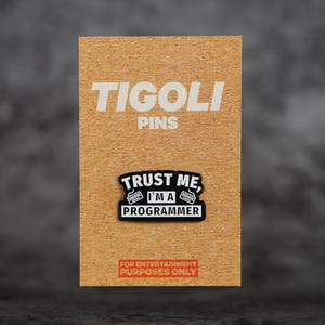 May include: A rectangular pin on a brown card. The pin reads "TRUST ME, I'M A PROGRAMMER" in white text on a black background. The card also has the text "TIGOLI PINS" and "FOR ENTERTAINMENT PURPOSES ONLY".