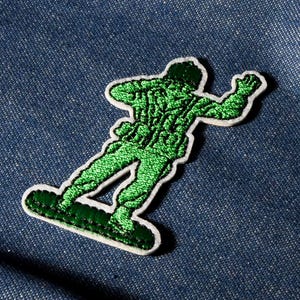 May include: A green embroidered patch of a toy soldier with a white border, set against a blue denim backdrop. The soldier is depicted in a dynamic pose, with one arm raised in the air.
