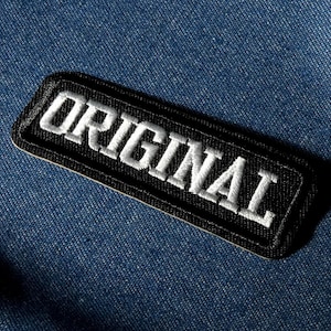May include: A black rectangular patch with the word "ORIGINAL" embroidered in white. The patch is set against a blue denim background. The white text stands out against the dark patch.