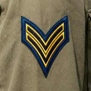 Op de afbeelding: Detail van een militair uniform. De olijfgroene stof toont een blauw en geel chevron-embleem, dat de rang aangeeft. Het embleem is geborduurd met een blauwe rand en twee gele strepen.
