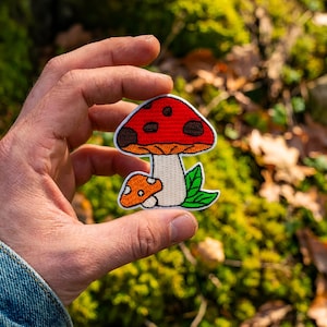 Può includere: Toppa ricamata a forma di fungo, con cappello rosso a pois marroni, gambo arancione e foglie verdi. Include anche un fungo arancione più piccolo. Perfetta per personalizzare abbigliamento o accessori.